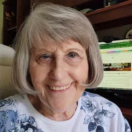 Picture of woman with short light blonde/grey hair wearing a white t-shirt and white cardigan sweater with blue flowers